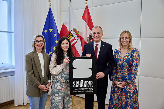 Portraitfoto von drei Frauen und einem Mann, halten ein Schild mit SCHÄXPIR, Fahnen im Hintergrund