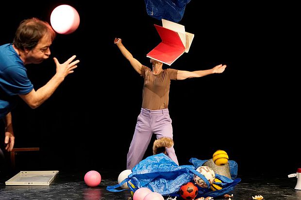 Eine Frau und ein Mann auf der Bühne tanzend, Bälle am Boden, ein Buch fliegt in der Luft