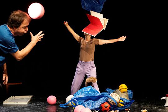 Eine Frau und ein Mann auf der Bühne tanzend, Bälle am Boden, ein Buch fliegt in der Luft